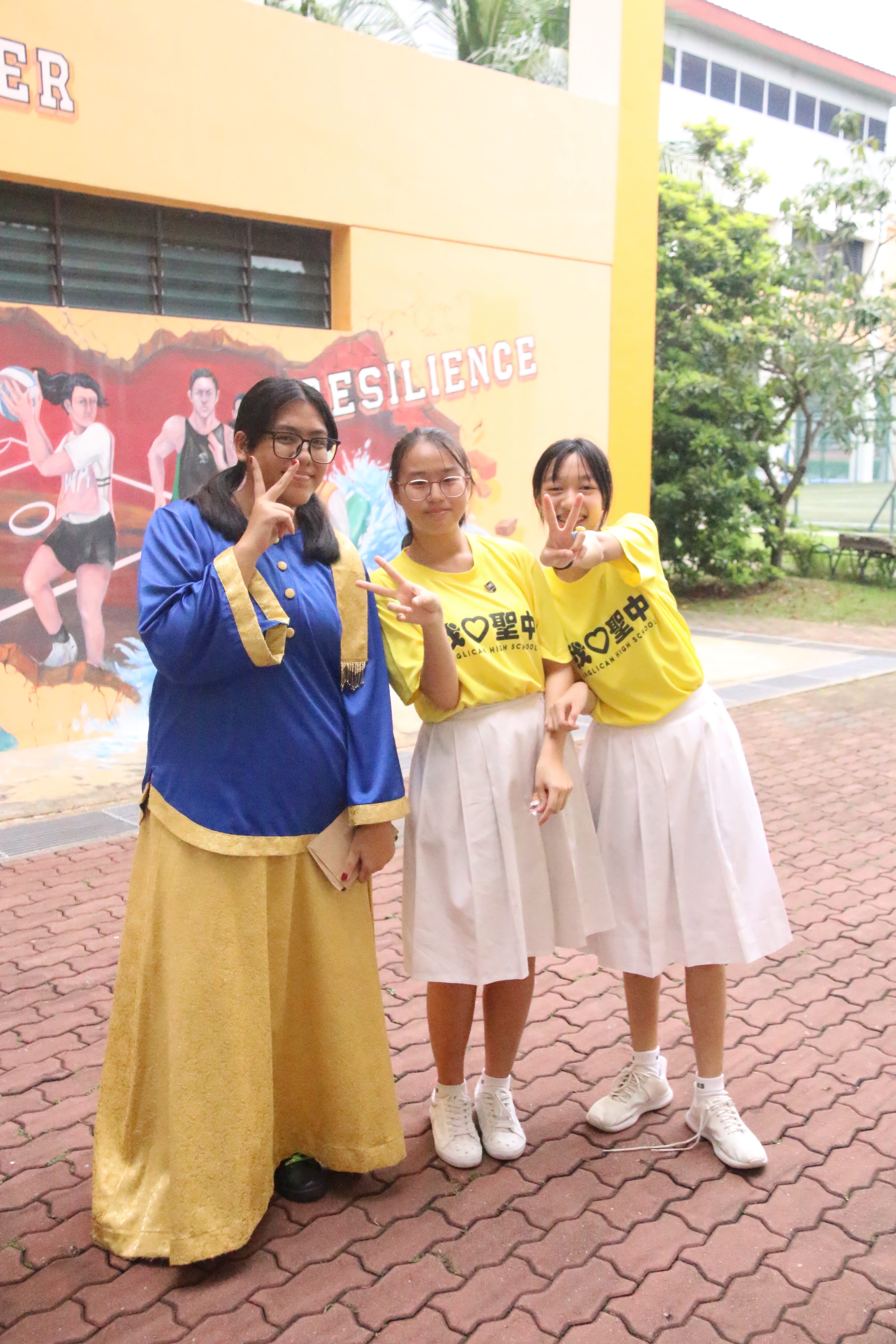 Three girls posing with peace signs outside a school, one in blue traditional dress, two in yellow school shirts.